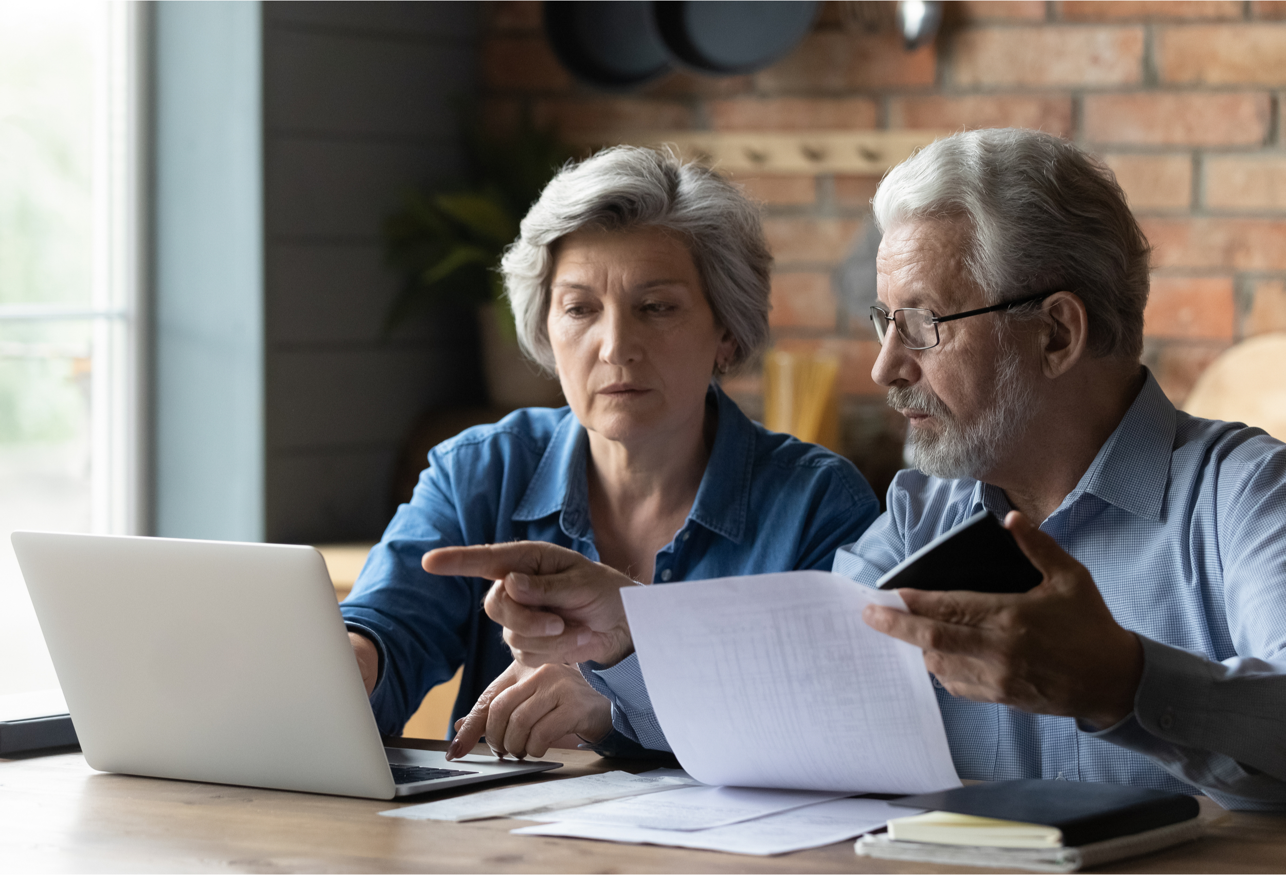 An older couple sitting at their kitchen table looking at a laptop and paperwork
