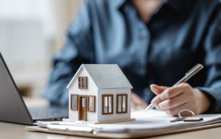 A person signing a document with a model house on the table.